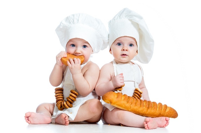 Dos niños pequeños blancos vestidos como chefs y sentados comiendo pan